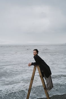 Elegant woman standing on a ladder by the ocean in a trench coat on a cloudy day.