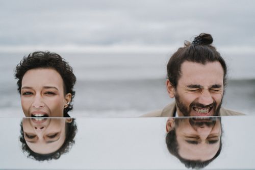 Free A joyful couple shares a laugh at the beach, captured with their reflection. Stock Photo