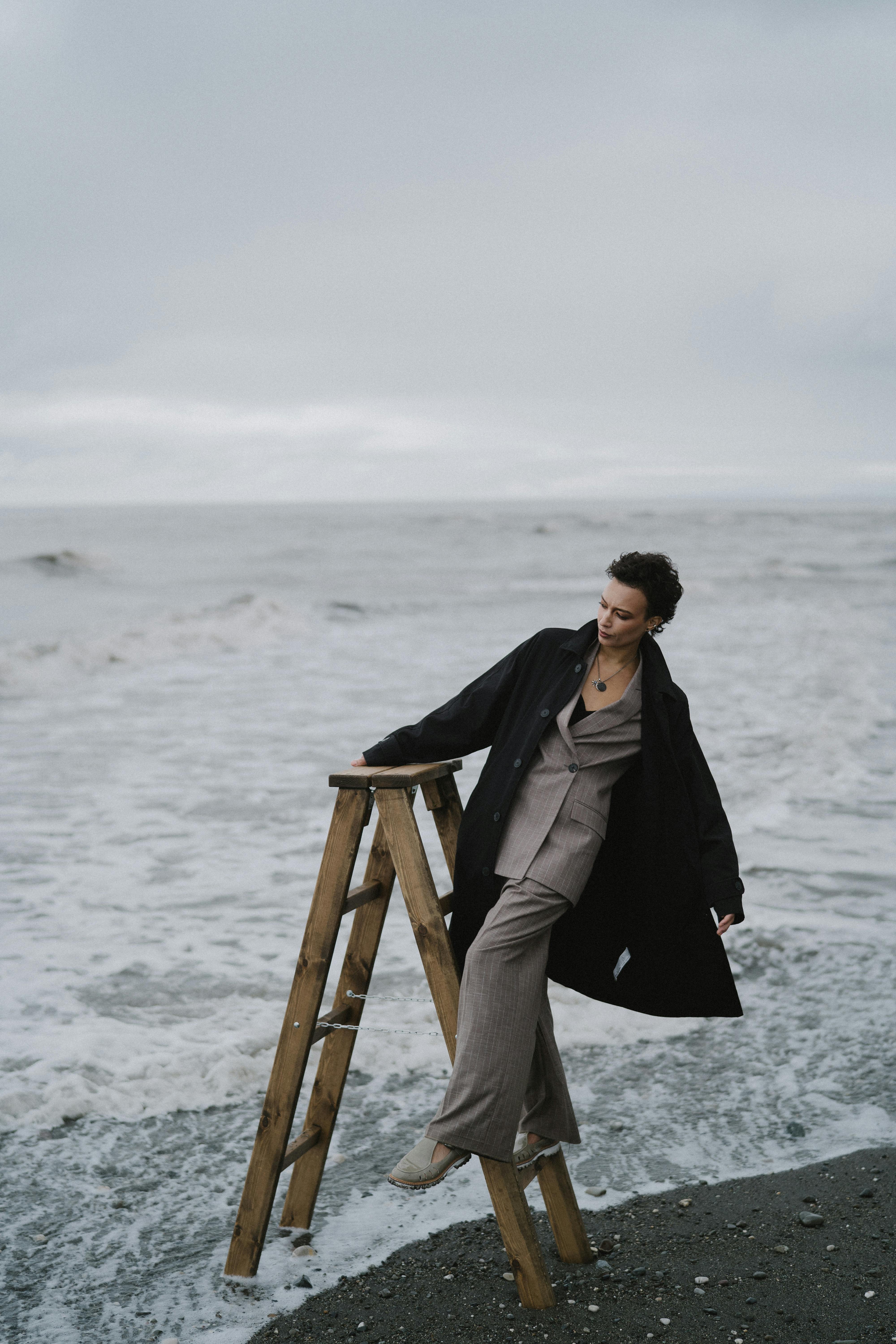 woman in black coat sitting on brown wooden chair on beach