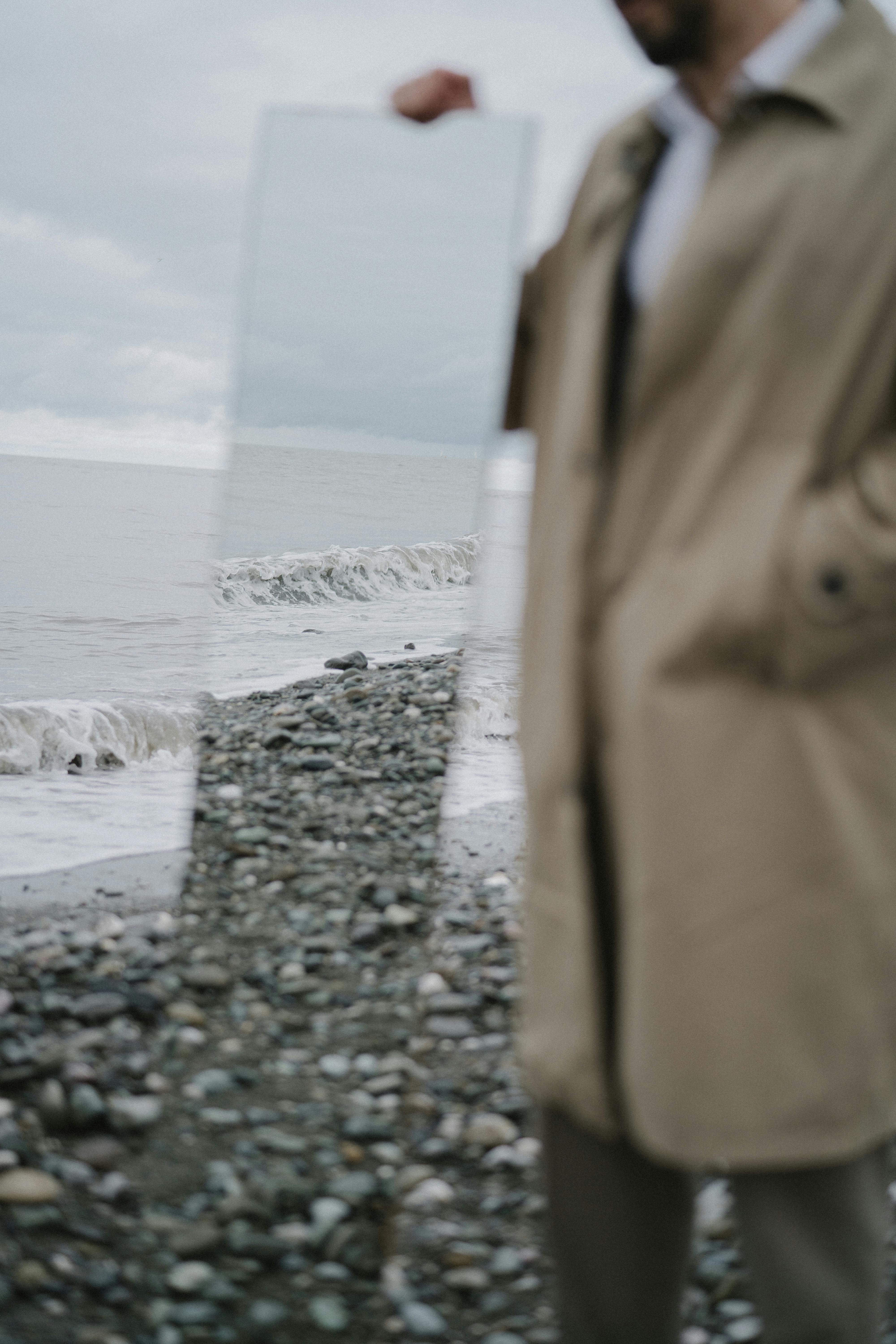 Person holding mirror on rocky beach reflecting the ocean waves.