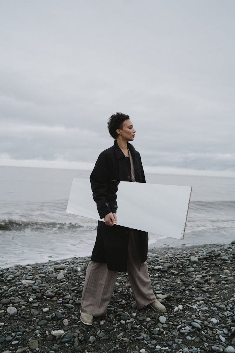 Woman In Black Trench Coat Holding A Mirror And Standing On A Seashore