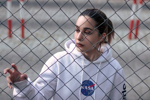 A woman in a white hoodie stands pensively behind a chain-link fence outdoors.