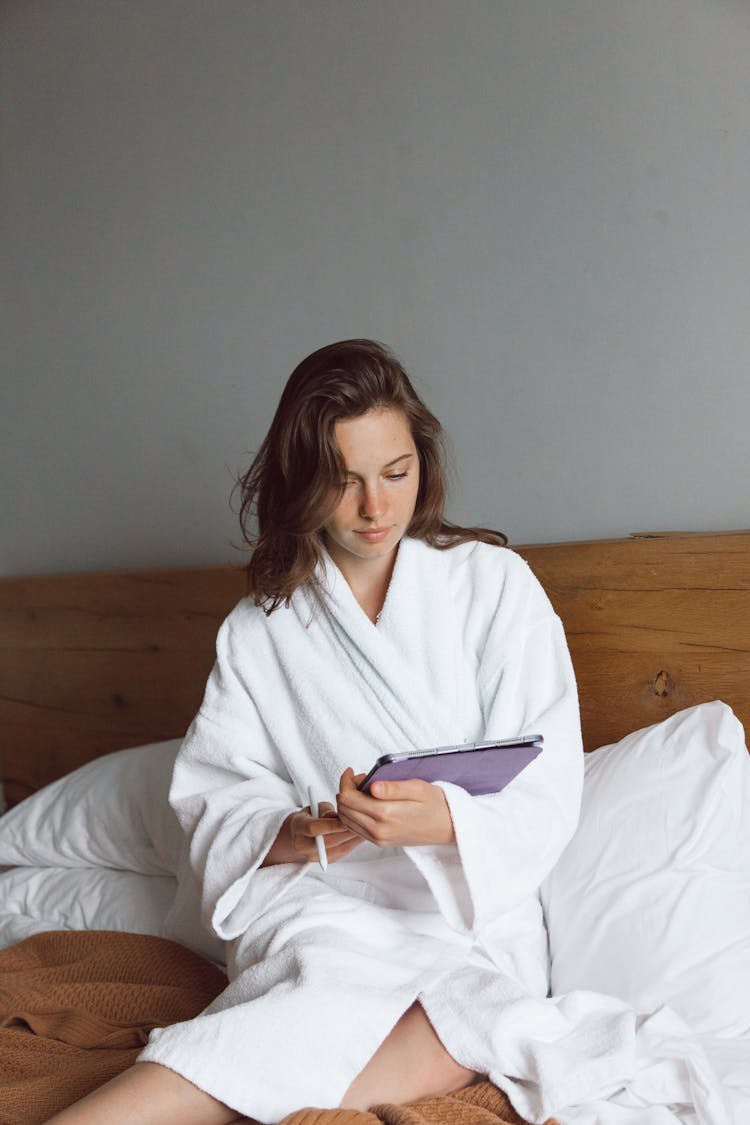 A Woman In White Bathrobe Sitting On The Bed While Holding A Tablet