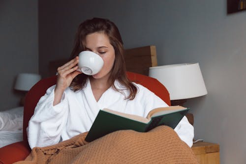 Free Woman in White Robe Drinking on White Ceramic Mug while Reading a Book Stock Photo