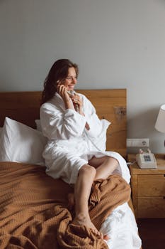 Smiling woman in bathrobe sits on bed, talking on a vintage telephone indoors.