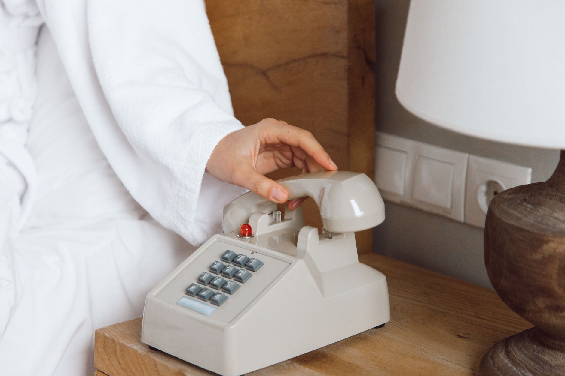 Person in white robe using a vintage rotary phone on a wooden nightstand, beside a lamp. Related image for Smart Hotels: How Brands Can Improve Performance.