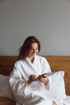 Smiling woman in white bathrobe using tablet while sitting on bed indoors.