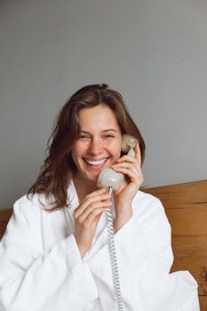 A cheerful woman in a bathrobe enjoys a conversation on a rotary phone indoors.