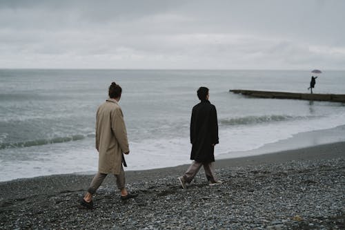 Couple Walking on Rocky Shore Under Gray Sky