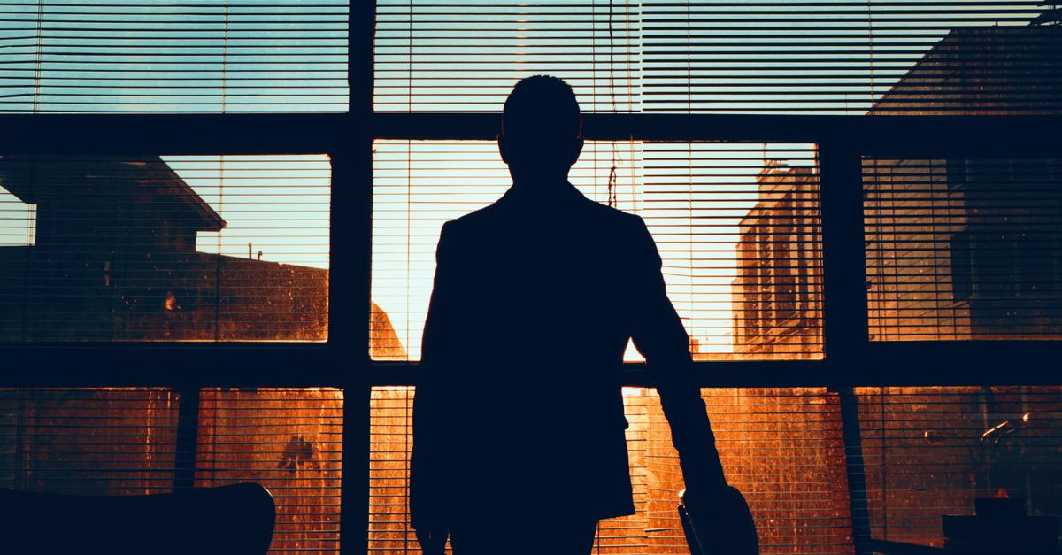 Silhouette of a man standing by a window, observing a building at sunset.