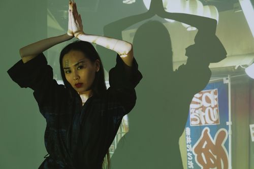 Free An artistic composition of an Asian woman posing with her hands raised, creating a shadow on the wall. Stock Photo