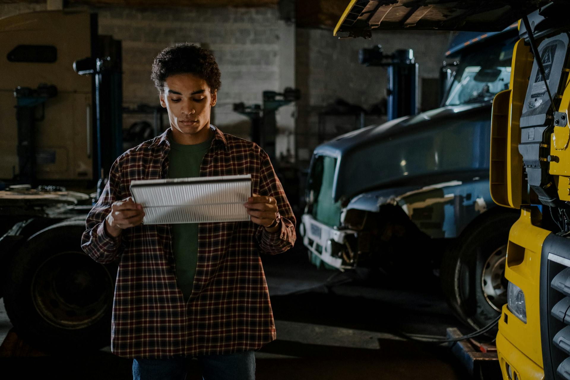 An auto mechanic inspects an air filter inside a dimly lit garage with trucks.