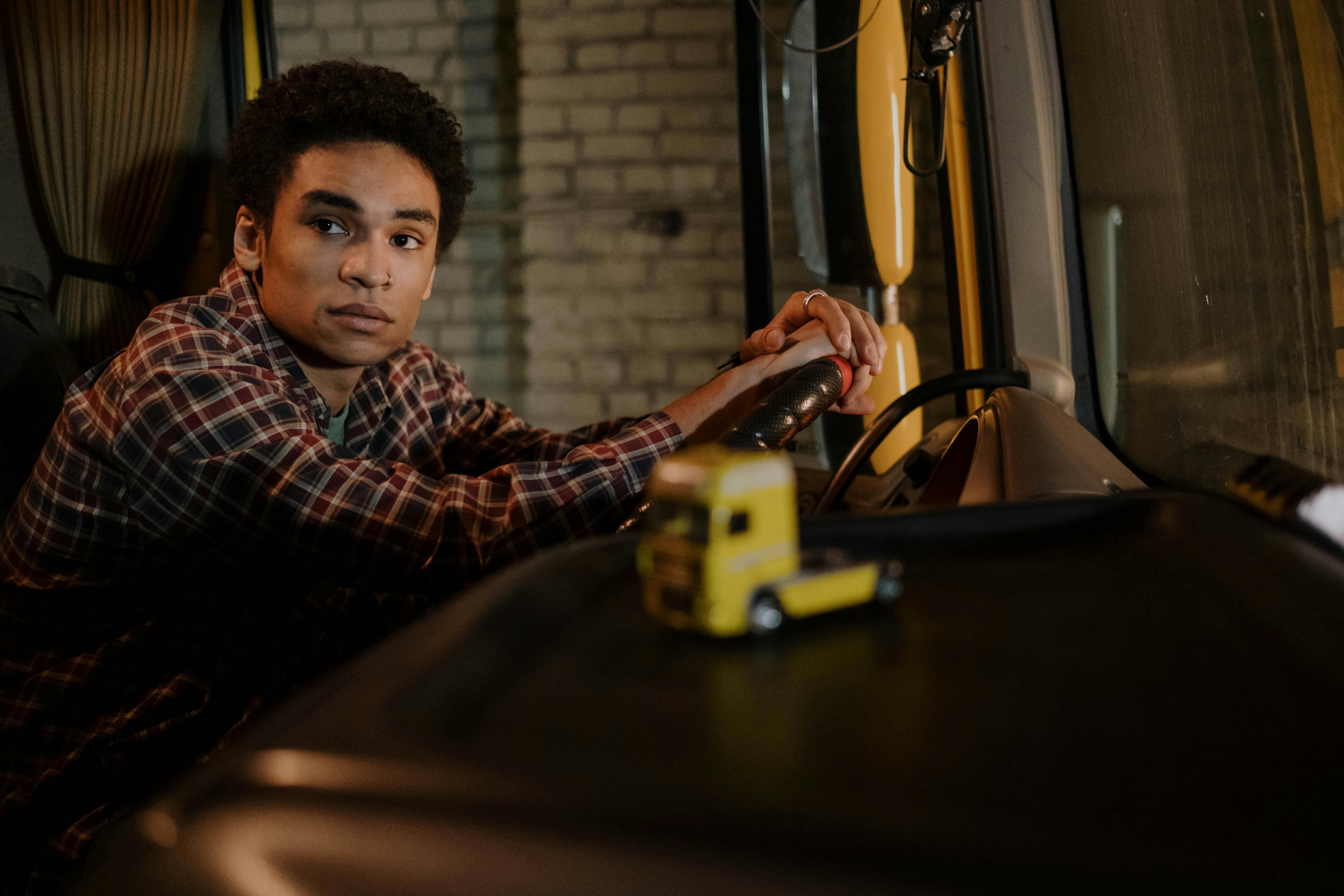 A young man sits in the cabin of a truck, hands on the steering wheel, with a model truck on the dashboard.