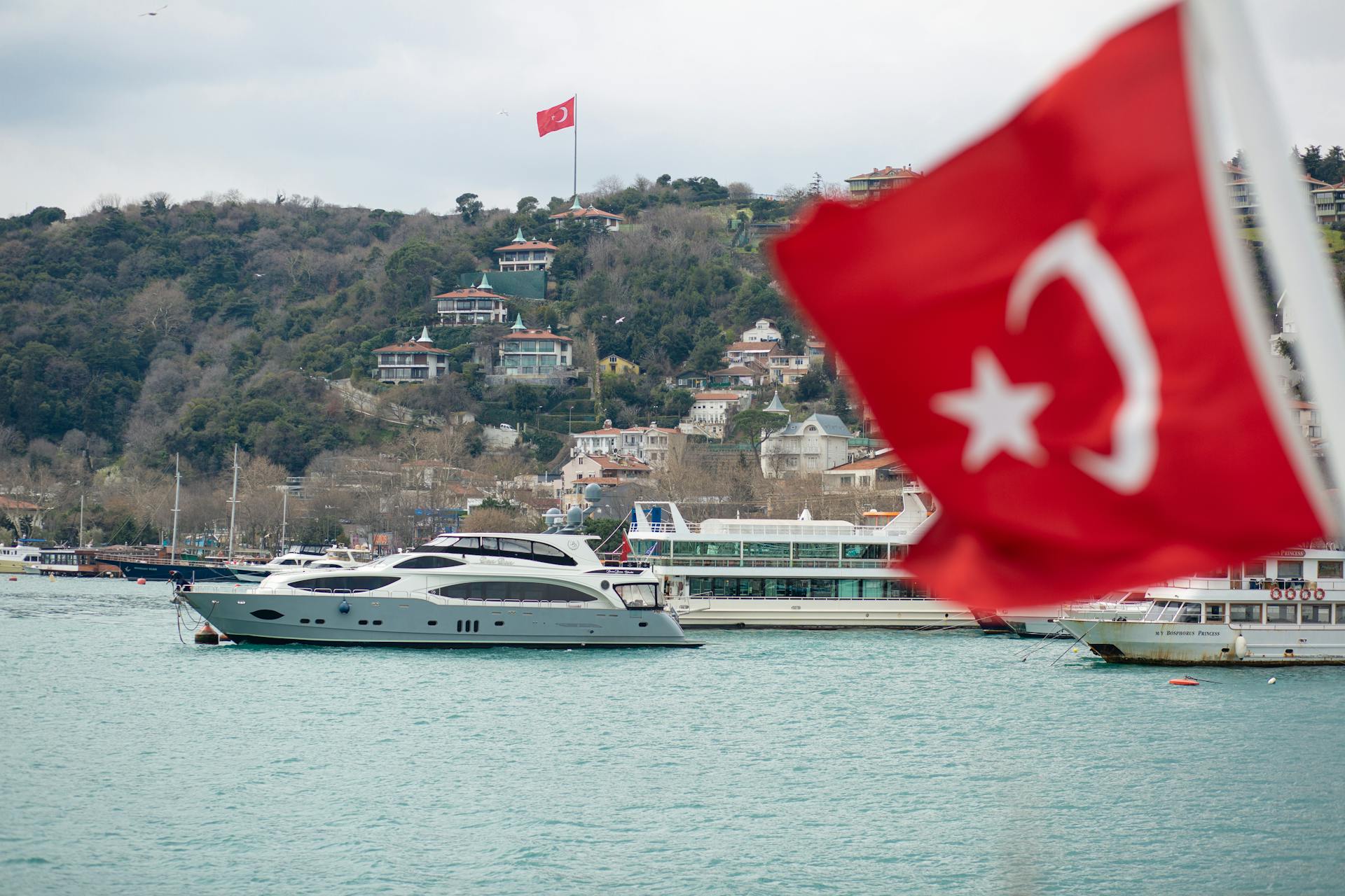 Modern yachts and excursion boats floating on rippling river near residential houses built on hill slope on cloudy day in Istanbul