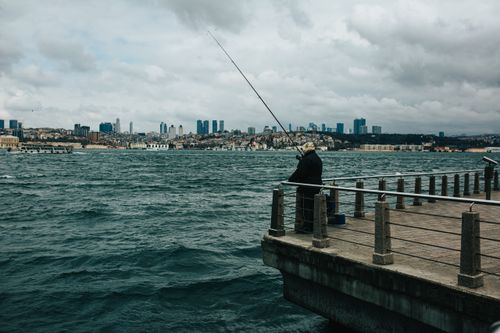 Free Back view of unrecognizable male in outerwear with fishing rod standing on pier against rippling river under cloudy sky Stock Photo
