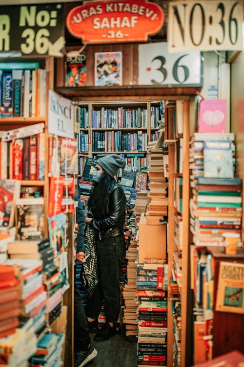 Free Side view full body of young female in warm clothes and mask standing in shop with large collection of various books Stock Photo