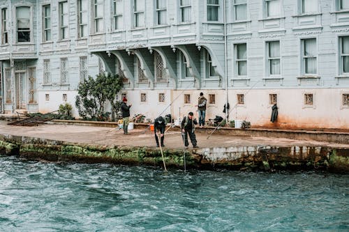 Fishermen in casual clothes standing and fishing on embankment against building in city in daytime