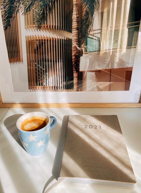 A cup of coffee next to a 2021 planner in sunlight with a framed art piece.