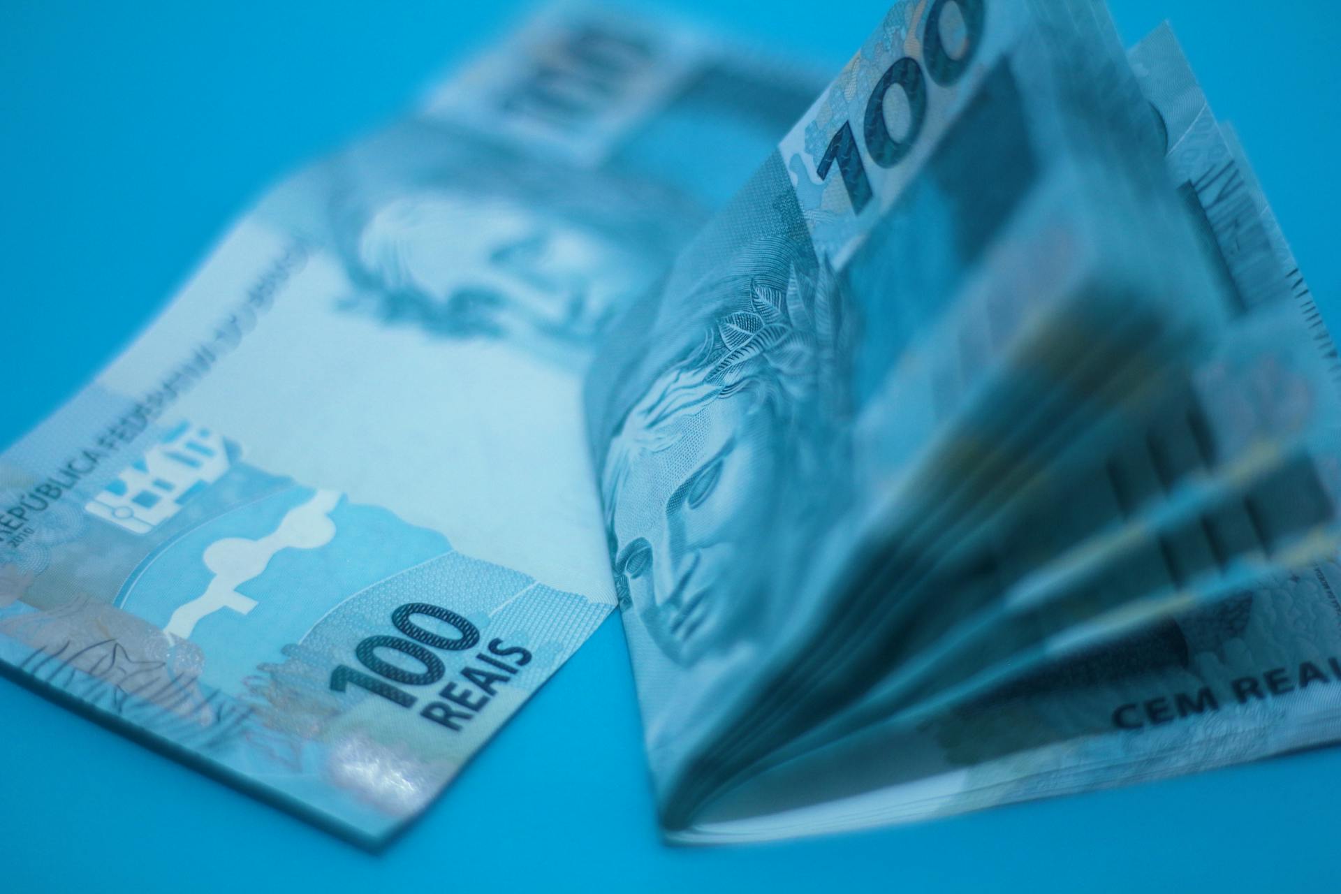 Stack of 100 Brazilian real notes placed on a blue surface, showcasing currency details.