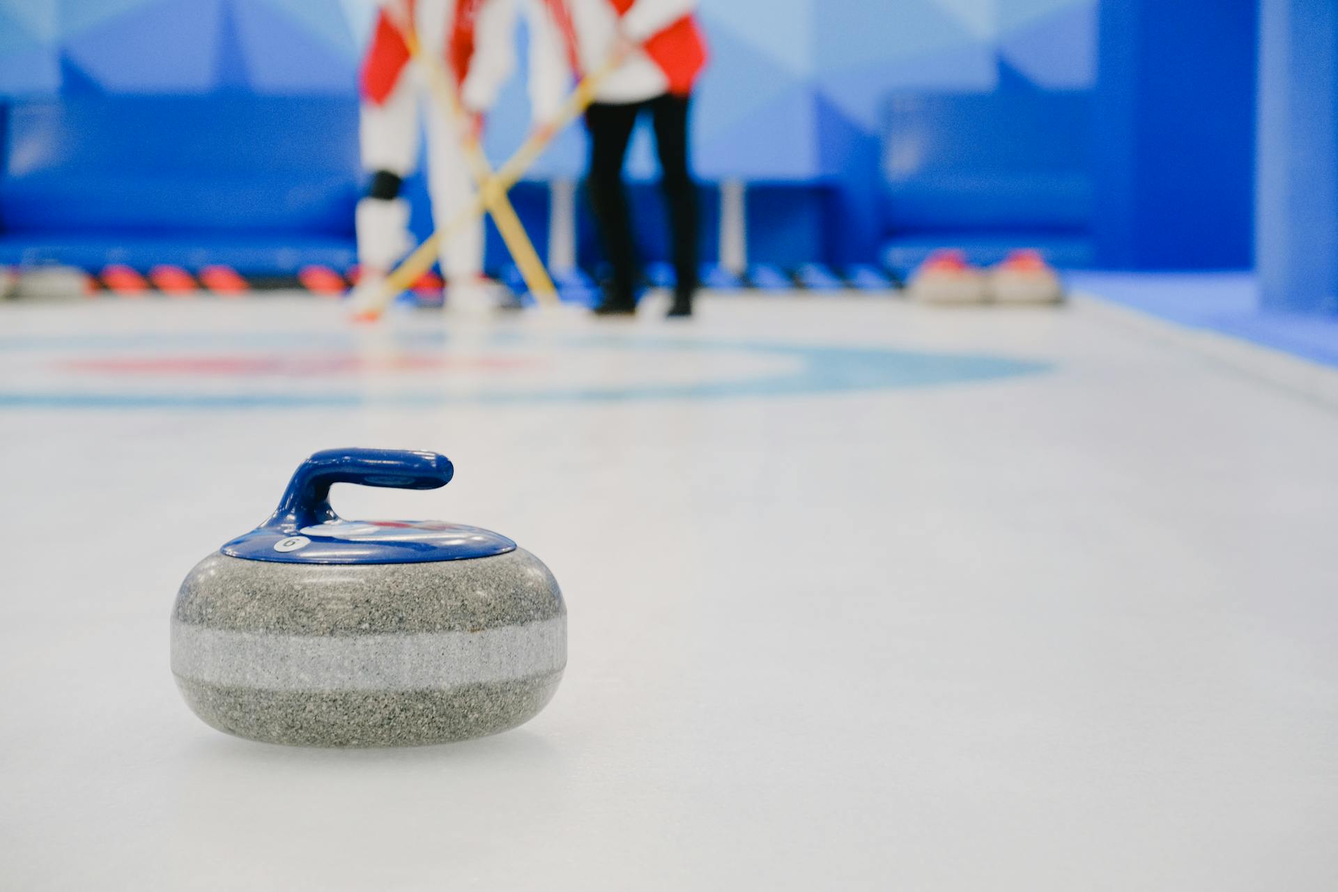 Curling stone on ice with blurred players sweeping in the background inside a rink.