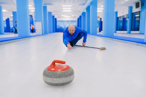 Focused sportsman in blue activewear standing on knee with broom stick and looking at sliding stone during curling training on ice rink