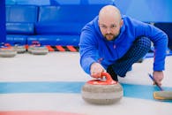 Concentrated sportsman in blue activewear squatting down and throwing heavy stone while playing curling on modern ice rink