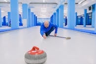 Concentrated sportsman with beard in blue uniform squatting down and looking at sliding stone with red handle during curling game on modern ice rink