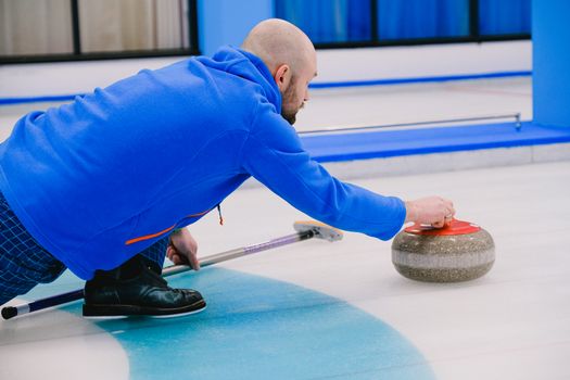 Side view of concentrated sportsman in sportswear sliding gray stone with red handle while playing curling on ice rink