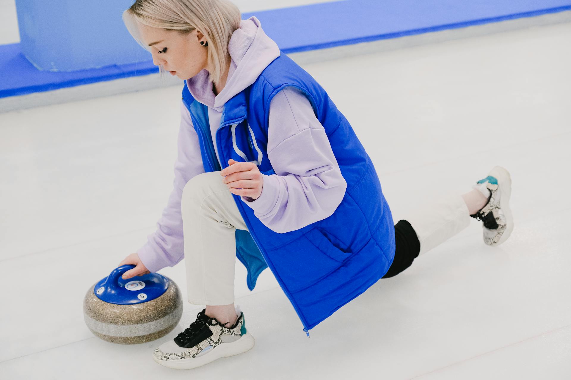 Full body of concentrated young female player wearing sport vest and sneakers standing on knee while moving blue handled stone while playing curling on ice arena