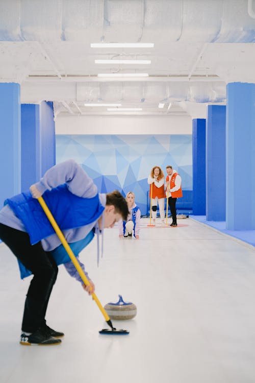 Young male player wearing warm vest jumper and black trousers sweeping ice with broom while playing curling in ice rink