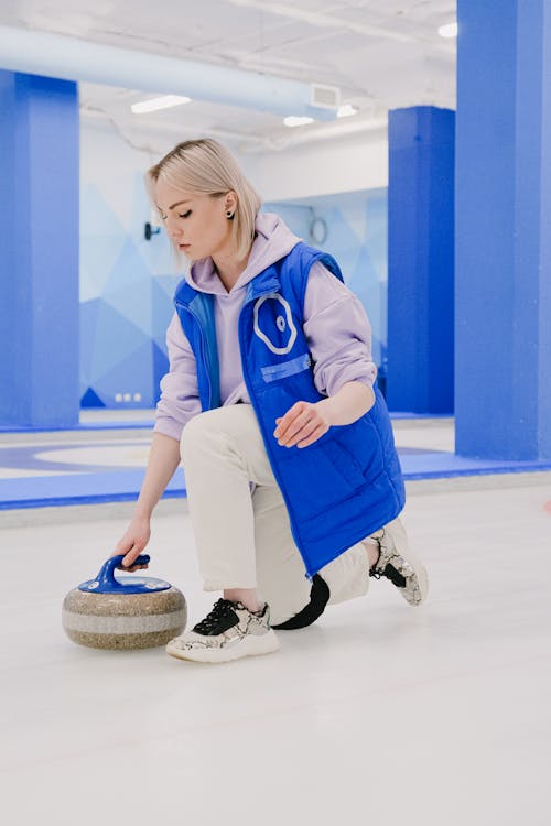 Full body side view of focused sportswoman in sportswear preparing to slide granite curling stone on ice sheet during sportive game