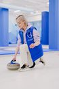 Full body side view of focused sportswoman in sportswear preparing to slide granite curling stone on ice sheet during sportive game