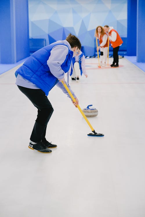 Unrecognizable sportsman in activewear sweeping ice sheet with broom in front of sliding curling stone while playing sportive game near team