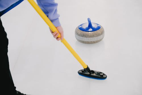 Crop anonymous sportsman with special broom sweeping ice sheet in front of sliding curling granite stone while playing sportive game