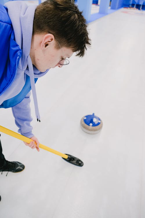 Side view of determined sportsman in sportswear with special broom in hands sweeping slippery ice in front of sliding curling stone during game