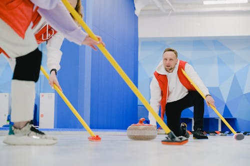 Determined sportsman throwing heavy granite curling stone near crop anonymous team sweeping ice sheet with brooms while playing sportive game