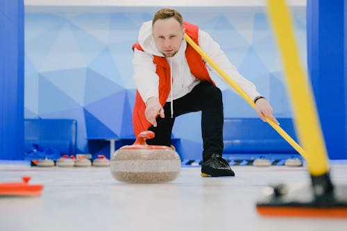 Determined sportsman in sportswear with special broom in hand throwing granite curling stone sliding on ice sheet while playing game