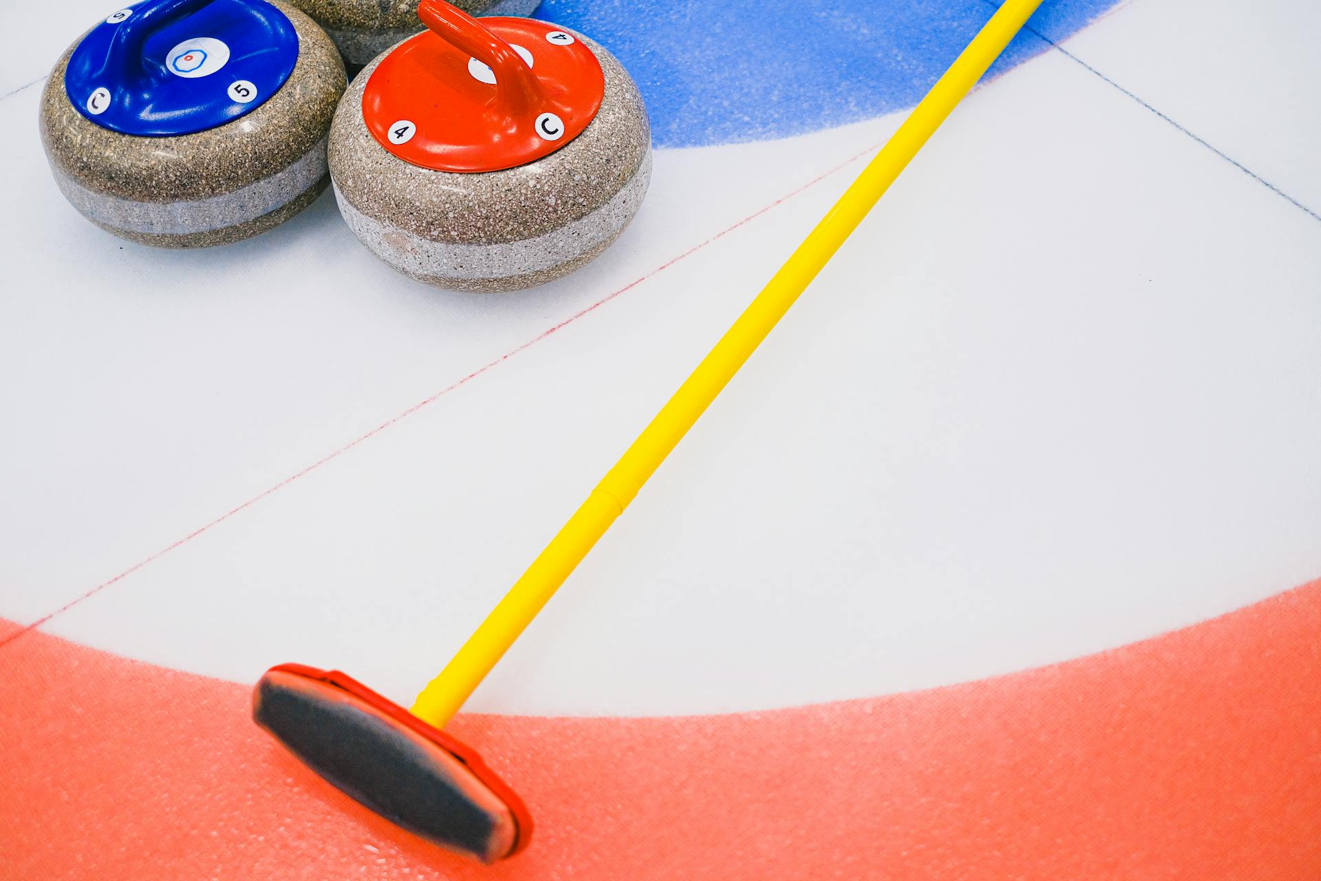 From above of granite curling stone and broom placed on ice sheet in house with colorful target area in playing area