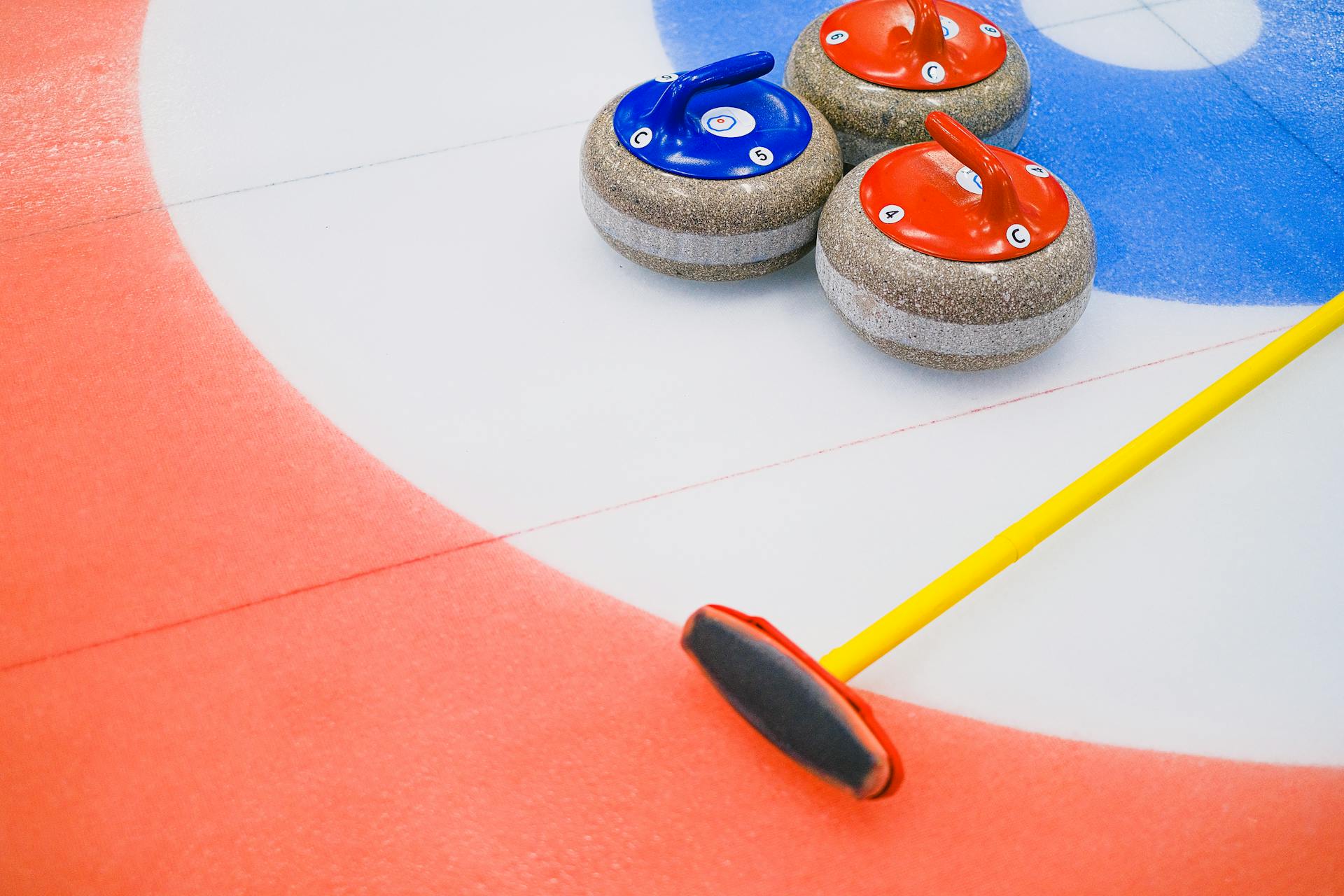 From above of heavy multicolored granite curling stones and long brush placed on ice with circular target area in arena