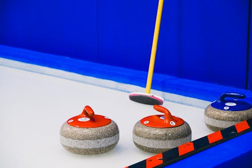 Curling stones with colorful handles and broom placed on ice sheet near blue wall and border during game in arena