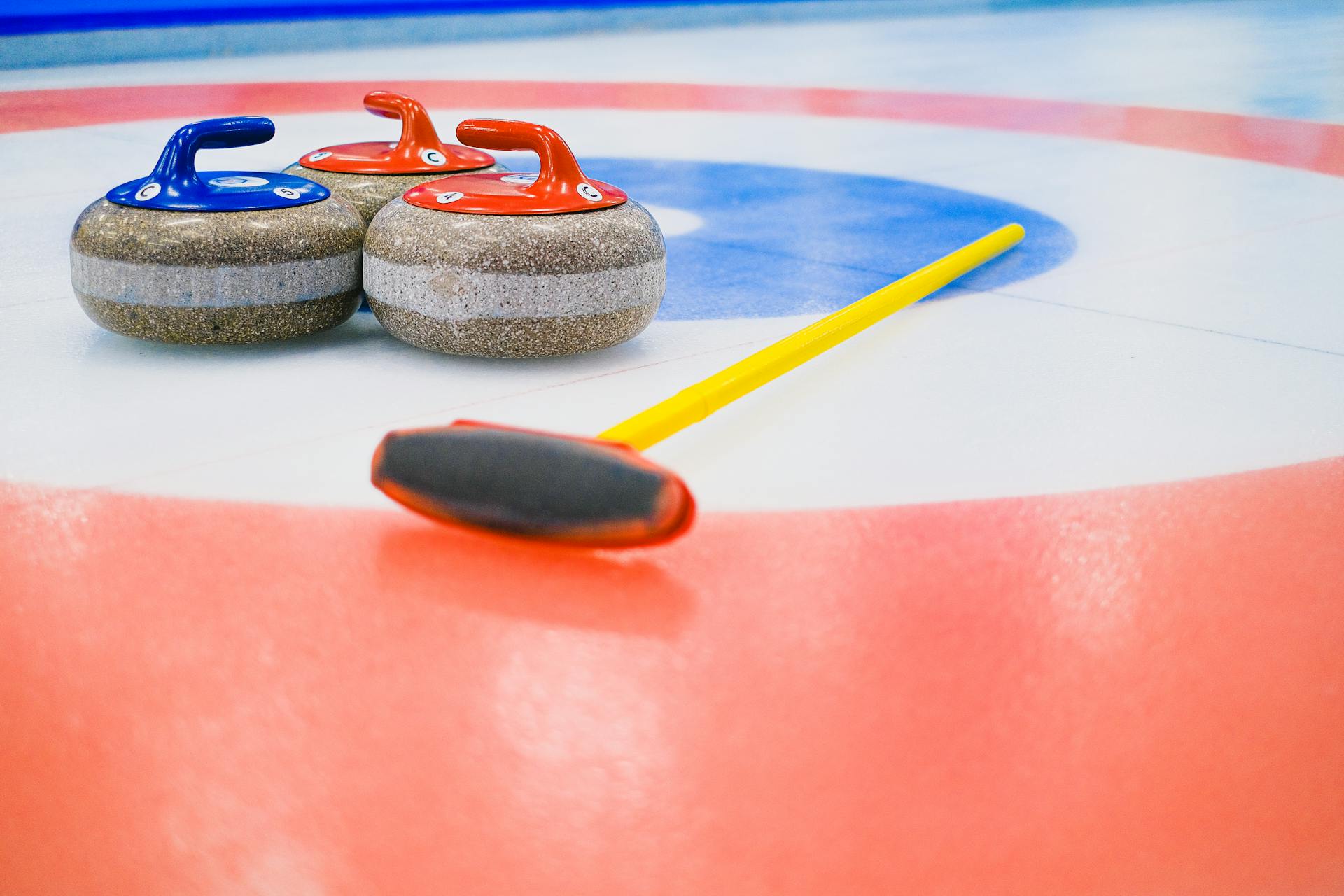 Heavy granite curling stones with handles and broom placed on ice in colorful circular target area during game in arena