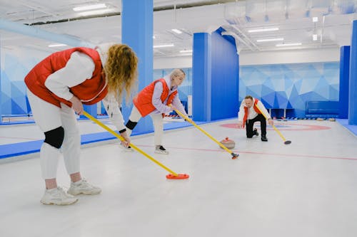 Full body of sportsman throwing curling stone near sportswomen in activewear sweeping ice sheet with special brooms while playing game