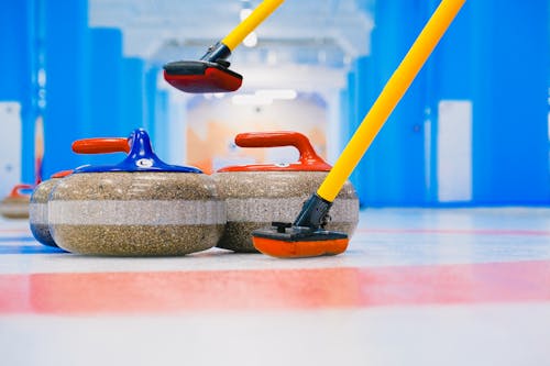 Granite curling stones with handles and long brooms placed on circular target area on ice sheet in arena with blue columns