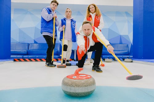 Sportsman throwing curling stone on ice sheet near team in sportswear with special brooms looking at camera while playing game