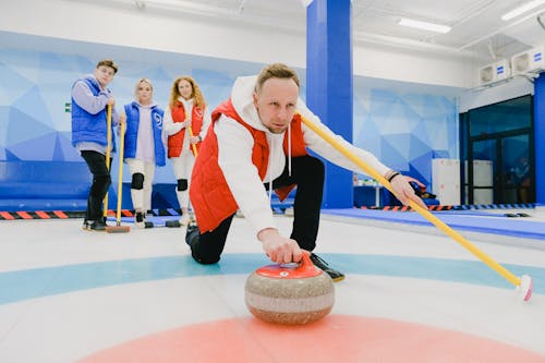 Serious man in sportswear with broom in hand preparing to throw granite curling stone on ice sheet near team during game