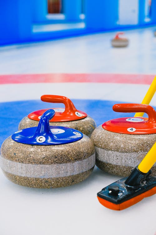 Colorful heavy curling stones with handles and broom placed in house with target area on slippery ice in arena during game