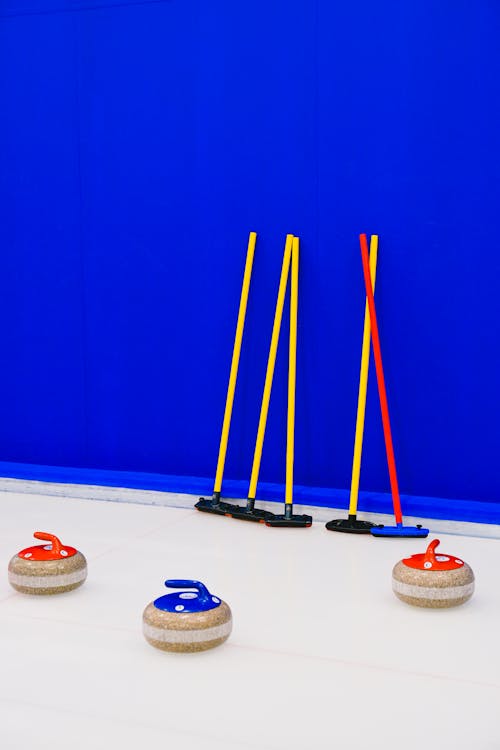 Colorful granite curling stones and set of broom placed near blue wall on clear ice sheet during game in arena