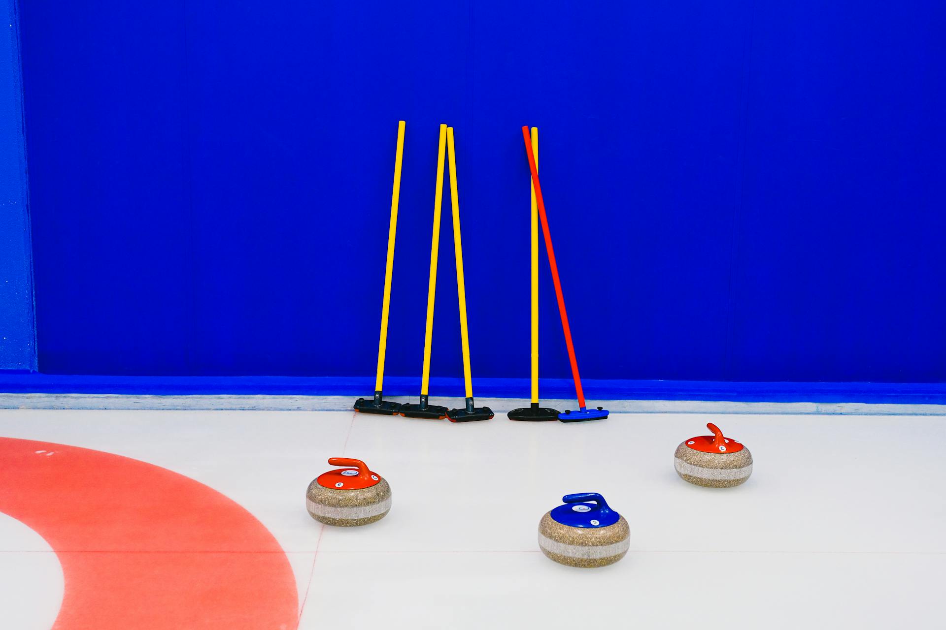 Granite curling stones with colorful handles placed on ice sheet near set of shrubs at blue wall in arena during game