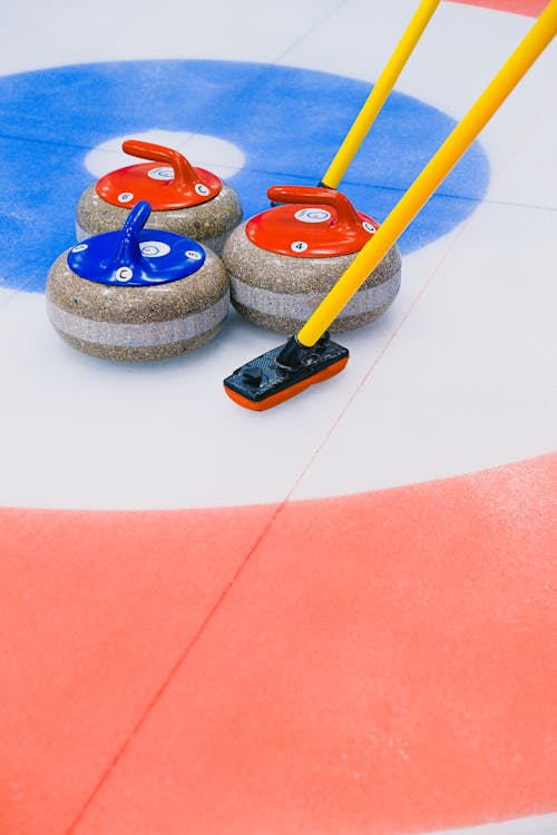 Granite curling stones with multicolored handles and brooms placed on ice sheet in circular target area in arena during game