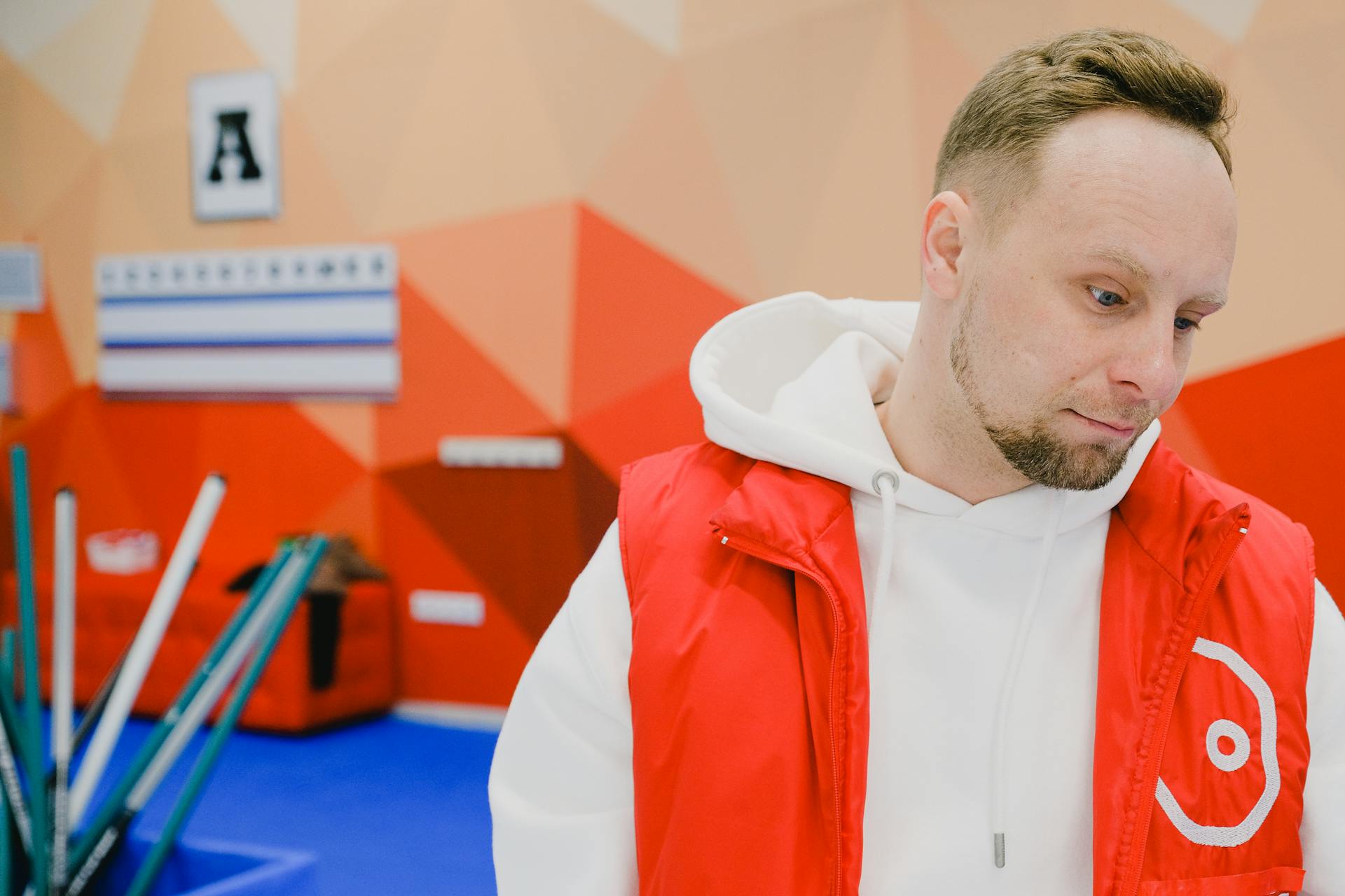 Pensive man in sportswear looking down while standing near curling brushes and wall with score board during game in arena